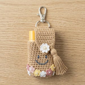 May include: A tan crochet holder for a small bottle, featuring a silver metal lobster clasp, a dark blue embroidered smiley face, a white flower accent on the flap, and a row of small pink, yellow, and white flowers with pearl centers.