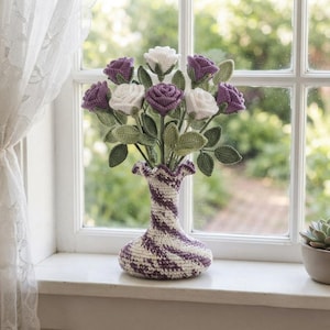 May include: A crochet vase with a purple and white marbled pattern holds a bouquet of handmade crochet roses. The flowers are in shades of deep purple and white, featuring green crochet leaves and stems, arranged in a ruffled-top yarn vase.