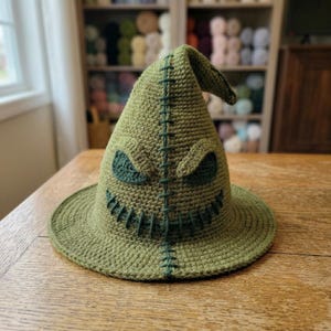 May include: A crocheted hat in olive green yarn, featuring a pointed, slightly slumped top and a wide brim. The hat displays dark green stitched details forming two triangular eyes and a wide, jagged mouth, with a vertical seam running down the front.