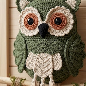 May include: A crochet owl wall hanging featuring a dark green body with textured wing patterns, large cream-colored scalloped eye patches, brown iris rings, black pupils, a black beak, and a cream-colored chest piece with three hanging tassels.