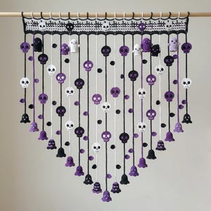 May include: A crochet wall hanging suspended from a wooden rod, featuring a black and white lattice header with small skulls. Multiple vertical strands hang down, decorated with alternating white, black, and purple skulls, ghosts, and bell shapes.
