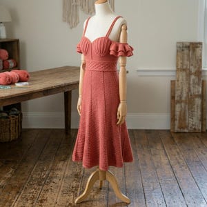 May include: A coral-colored, knee-length knit dress displayed on a mannequin. The dress features a sweetheart neckline, thin shoulder straps, and off-the-shoulder ruffled sleeves. The bodice includes a textured, crisscross pattern at the waist.