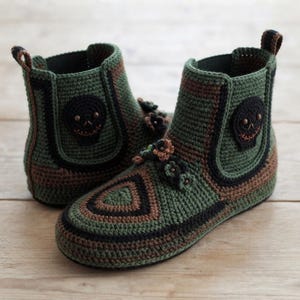 May include: A pair of ankle-high crochet boots in olive green, brown, and black yarn. Each boot features a black circular skull patch on the side, a pull tab at the heel, and a cluster of small crocheted flowers on the top of the foot area.