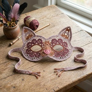 May include: A cat-shaped crochet mask in shades of dusty rose, mauve, and copper. The mask features intricate lace-like stitching, pointed ears, and a small heart-shaped nose detail. Two long crochet ties extend from the sides, finished with small beads.