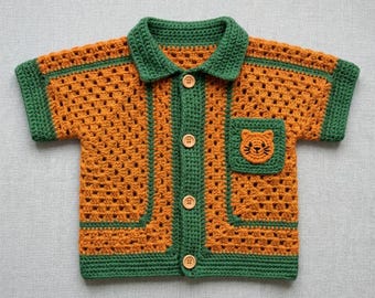 May include: A short-sleeved crochet shirt featuring a granny square pattern in orange with dark green trim on the collar, sleeves, and button placket. The shirt has four light brown buttons and a square chest pocket with an orange cat face design.