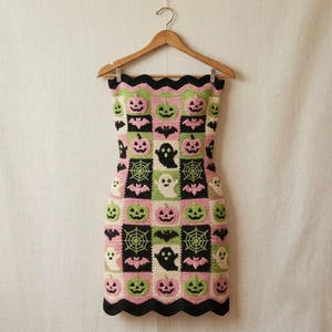 May include: A strapless crochet dress featuring a grid pattern of squares in black, light green, and light pink. The squares display motifs of jack-o'-lanterns, ghosts, bats, and spiderwebs. The dress has a scalloped top and bottom edge.