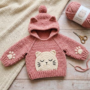 May include: A handmade pink crochet hoodie for a baby or toddler, featuring cat ears on the hood and a cat face applique on the front. The sleeves have paw print details. A ball of pink yarn and scissors are next to the hoodie.