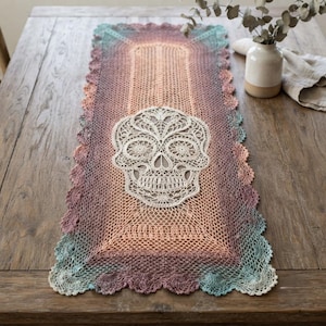 May include: A rectangular crochet table runner featuring a gradient color transition from teal to purple and orange. The center displays an intricate, cream-colored lace skull motif. The runner has a scalloped edge and a fine, open-work mesh pattern.