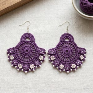 Può includere: Un paio di orecchini all'uncinetto viola con un design circolare a ventaglio e punti complessi. Gli orecchini sono decorati con piccoli fiori all'uncinetto bianchi e viola chiaro e minuscole perline bianche lungo il bordo inferiore.