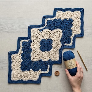 May include: A stepped, geometric crochet runner featuring alternating dark blue and cream-colored yarn in a repeating square motif with intricate lace-like edges. A ball of dark blue Artisan Lace Yarn and a gold crochet hook are positioned nearby.