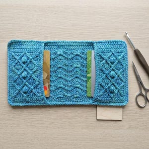 May include: A handmade, open turquoise crochet wallet with a diamond and wave pattern. The wallet is open, revealing two cards. A crochet hook and small scissors are next to the wallet on a light wood surface.