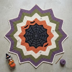 May include: A star-shaped crochet mat featuring a central black spiderweb pattern surrounded by concentric rings of orange, white, olive green, and purple yarn. The design includes small black bat and white ghost motifs integrated into the fabric.