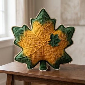 May include: A maple leaf-shaped crochet pillow featuring a gradient of yellow and green yarn. The edges are trimmed with small white beads, and the surface is decorated with two smaller, separate crochet leaf appliqués in yellow and green.