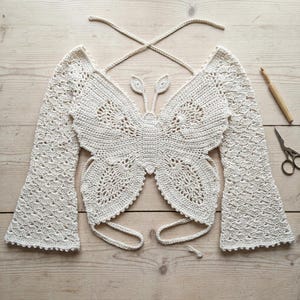 May include: An off-white crochet top shaped like a butterfly with long, flared sleeves and thin tie-back straps. The garment features intricate openwork patterns throughout the wings and body, accompanied by a wooden crochet hook and metal scissors.