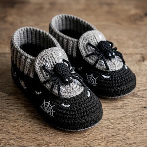 May include: A pair of black and gray crocheted booties featuring a textured spider on the top of each shoe. The black base is decorated with white embroidered spiderwebs and small bat motifs, while the gray ribbed cuffs provide contrast.