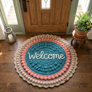 May include: A round, handmade crochet rug with a teal center, bordered by coral and tan sections. The word "Welcome" is spelled out in white script across the center. The rug is placed in front of a wooden door.