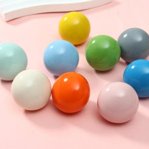 May include: A collection of nine smooth, spherical balls in various solid colors including yellow, light blue, green, gray, white, orange, pink, and teal. Each ball has a glossy finish and a uniform, round shape, arranged in a cluster.