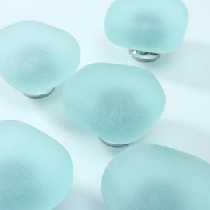 Puede incluir: Un conjunto de cinco pomos para armarios con tapas de vidrio esmerilado en color verde espuma de mar, lisas y redondeadas. Cada pomo está unido a una base metálica circular de tono plateado, ofreciendo un aspecto limpio y uniforme.