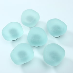 May include: A set of six cabinet knobs featuring rounded, frosted light blue glass tops with a smooth, matte texture. Each knob is attached to a circular metal base, providing a uniform appearance for furniture hardware or drawer pulls.