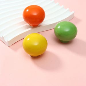 Puede incluir: Tres pomos esféricos para armarios con un acabado liso y brillante en naranja vibrante, amarillo brillante y verde claro. Cada pomo presenta una parte superior redondeada y una base pequeña, presentados como un conjunto de herrajes.