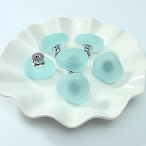 Puede incluir: Cinco pomos para armario de vidrio esmerilado en color azul claro, con formas redondeadas y lisas similares a guijarros. Cada pomo cuenta con una base de metal plateado pulido y un punto de fijación central, con un acabado mate translúcido.