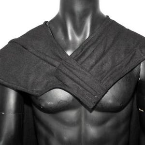 May include: A solid black fabric cape featuring a structured, overlapping wrap design across the chest and shoulders. The garment is draped over a dark, featureless torso mannequin, showcasing a clean, minimalist aesthetic with visible stitching.