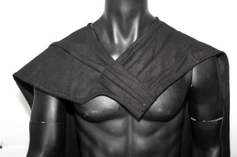 May include: A solid black fabric cape featuring a structured, overlapping wrap design across the chest and shoulders. The garment is draped over a dark, featureless torso mannequin, showcasing a clean, minimalist aesthetic with visible stitching.