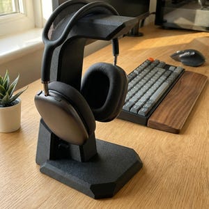 Può includere: Supporto nero per cuffie con cuffie nere. Il supporto ha un design moderno e una finitura strutturata. Le cuffie sono appese al supporto. Sullo sfondo sono visibili una tastiera e un mouse, che suggeriscono una configurazione dello spazio di lavoro.