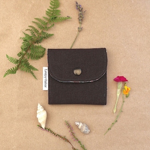 Op de afbeelding: Een vierkante donkerbruine stoffen portemonnee met een afgeronde flap en een enkele donkere knoopsluiting. Een wit stoffen label met de tekst Leepy Atelier is aan de zijkant bevestigd. De portemonnee is omringd door een varenblad, lavendel, bloemen en schelpen.