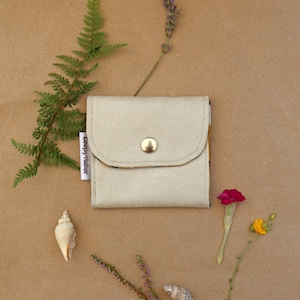 Op de afbeelding: Een vierkante, lichtbeige portemonnee met een afgeronde flap en een enkele goudkleurige metalen drukknoopsluiting. Een klein wit stoffen label met de tekst "LeepyFlutter" is aan de linkerkant van de portemonnee bevestigd, voorzien van zichtbare stiksels.