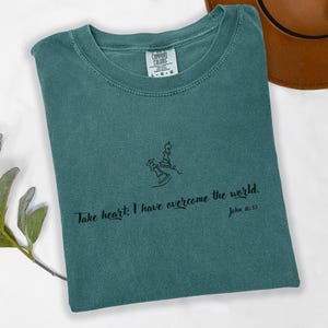 Może przedstawiać: Turkusowy t-shirt z krótkim rękawem i okrągłym dekoltem. Na środku koszulki znajduje się czarna grafika liniowa przedstawiająca dwie figury szachowe oraz napis "Take heart; I have overcome the world. John 16:33" wykonany kursywą.