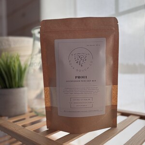 Peut inclure: Un sac en papier brun de mélange à crêpes au levain PROFI de Nova Dough Co. Le sac porte une étiquette blanche avec le logo de l'entreprise et des informations sur le produit. Le sac est posé sur une étagère en bois. Poids net : 410 g.