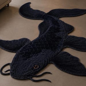 May include: A dark navy blue rug shaped like a koi fish. The rug features a textured surface with raised scale patterns along the body, detailed fin designs, and two long barbels extending from the mouth. A single visible eye has a brown iris.