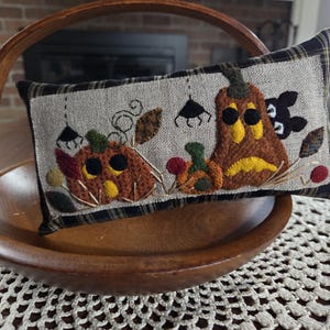May include: A rectangular fabric pillow featuring an embroidered scene of three pumpkins and a black cat on a light tan background. The pillow is bordered by a black and tan plaid fabric and displays decorative stitching of vines and small berries.