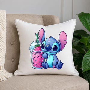 May include: A white square throw pillow featuring a colorful graphic of a blue alien character with large ears sitting next to a clear plastic cup filled with a pink strawberry drink, complete with a green straw and a strawberry garnish.