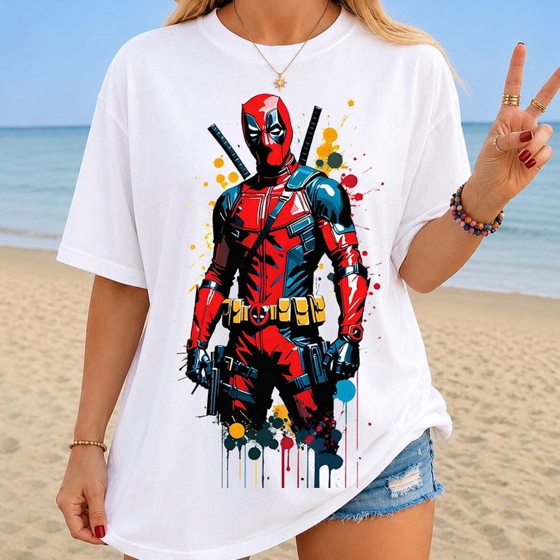 May include: A white short-sleeve t-shirt featuring a graphic print of the character Deadpool in a red and black suit with two swords on the back. The design includes colorful paint splatter accents in yellow, blue, and red around the character.