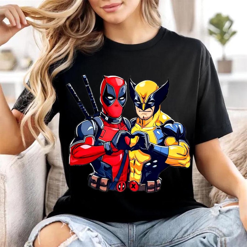 May include: A black short-sleeved t-shirt featuring a graphic print of the characters Deadpool and Wolverine standing side by side. Deadpool is in a red and black suit with swords on his back, while Wolverine wears a yellow and blue suit.