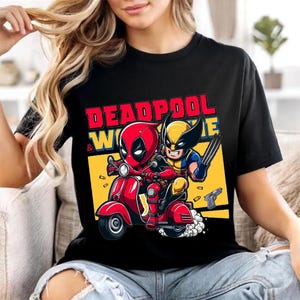 May include: A black t-shirt featuring a graphic print of Deadpool and Wolverine riding a red scooter. The text "DEADPOOL & WOLVERINE" is printed in red and yellow above the characters. The background includes a yellow rectangle and floating bullets.