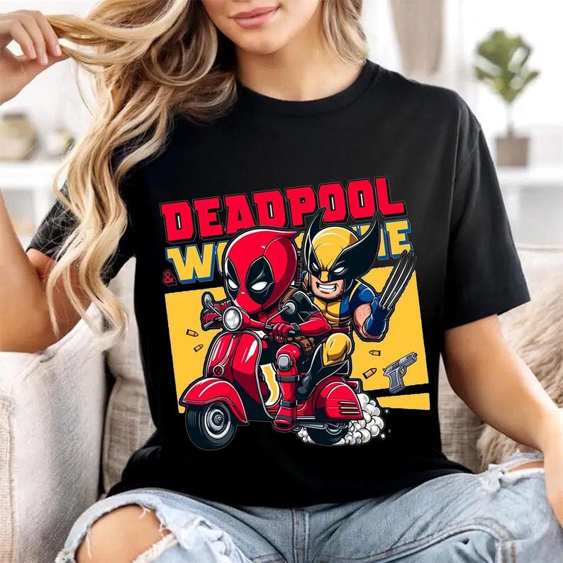 May include: A black t-shirt featuring a graphic print of Deadpool and Wolverine riding a red scooter. The text "DEADPOOL & WOLVERINE" is printed in red and yellow above the characters. The background includes a yellow rectangle and floating bullets.