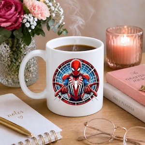 May include: A white ceramic mug featuring a circular graphic of the superhero Spider-Man in a red and blue suit with a white spider emblem on the chest, shown in a dynamic pose with hands extended forward against a blue webbed background.