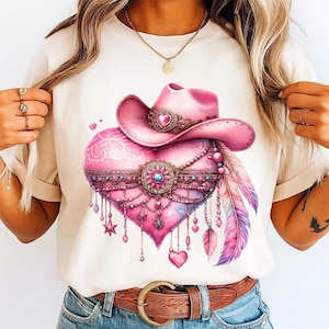 Pu&ograve; includere: Una t-shirt color crema con una stampa grafica di un cuore rosa decorato con un cappello da cowboy rosa, intricati filigrani metallici, catenelle pendenti, piccoli ciondoli a forma di stella e due grandi piume nei toni del rosa, viola e blu.