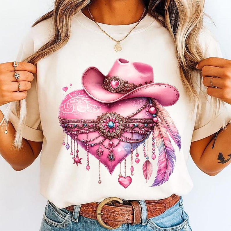 Pu&ograve; includere: Una t-shirt color crema con una stampa grafica di un cuore rosa decorato con un cappello da cowboy rosa, intricati filigrani metallici, catenelle pendenti, piccoli ciondoli a forma di stella e due grandi piume nei toni del rosa, viola e blu.