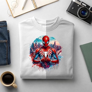 May include: A white crew neck t-shirt featuring a vibrant graphic of a red and blue superhero with a large white spider emblem on the chest, set against a detailed city skyline background with colorful paint splatter accents around the circular design.