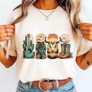 Pu&ograve; includere: Una t-shirt color crema con una stampa grafica raffigurante una chitarra acustica verde acqua accanto a un cactus, una persona con un cappello da cowboy, un teschio di bue e un paio di stivali da cowboy marroni con disegni di girasoli.