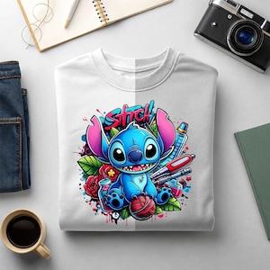 May include: A white crew neck t-shirt featuring a colorful graphic of the blue character Stitch from Lilo and Stitch. The design includes the word Stitch in graffiti-style lettering, red roses, a ball of yarn, and various art supplies.