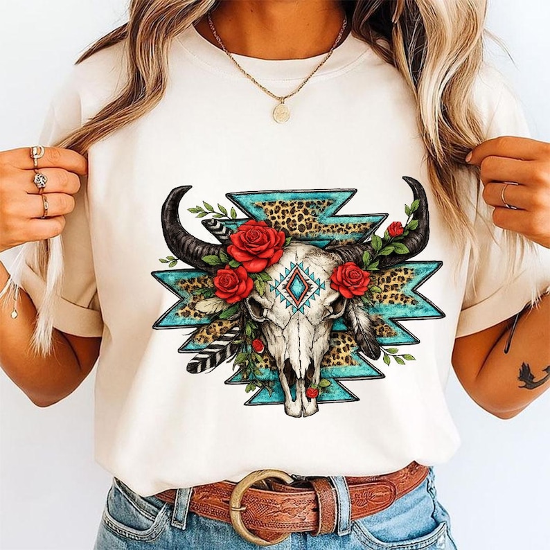 Pu&ograve; includere: Una t-shirt color crema con una stampa grafica di un teschio di toro decorato con rose rosse, foglie verdi e piume. Il design include una forma geometrica turchese, accenti con stampa leopardata e un motivo a rombi centrale.