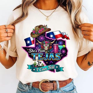 Pu&ograve; includere: Una t-shirt color crema con una grafica dello stato del Texas riempita con un motivo galattico, un teschio di mucca, un cappello da cowboy leopardato con fiori, un margarita, la bandiera del Texas e il testo She's From TEJAS I Can Tell.