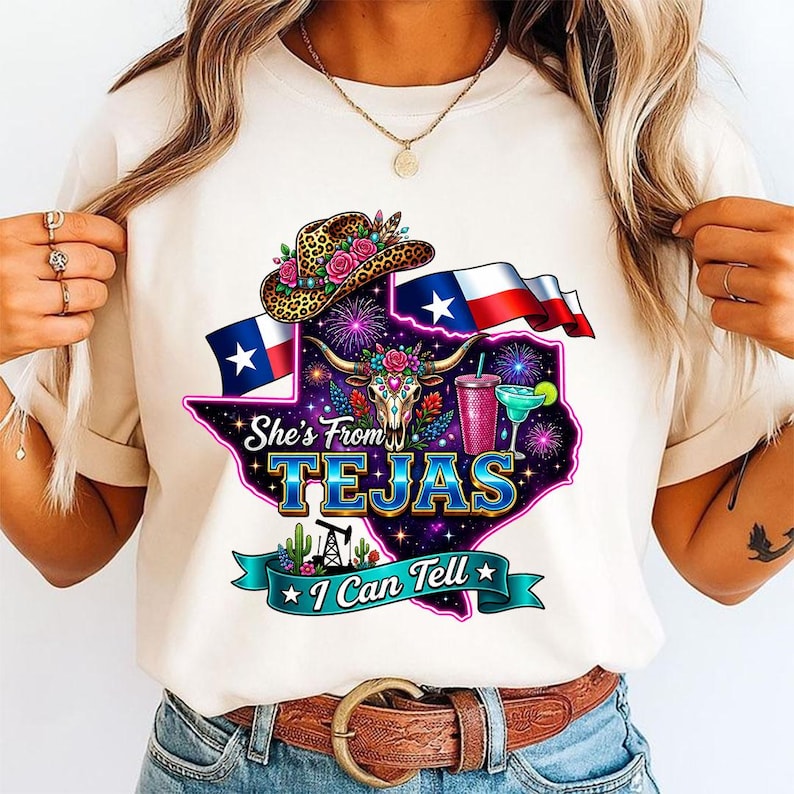 Pu&ograve; includere: Una t-shirt color crema con una grafica dello stato del Texas riempita con un motivo galattico, un teschio di mucca, un cappello da cowboy leopardato con fiori, un margarita, la bandiera del Texas e il testo She's From TEJAS I Can Tell.