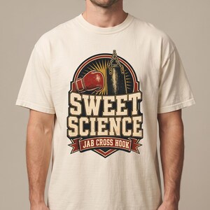 May include: A cream-colored short-sleeve t-shirt featuring a graphic print of a red boxing glove and a black punching bag. The text "SWEET SCIENCE" appears in large block letters above a banner that reads "JAB CROSS HOOK" in a dark red color.