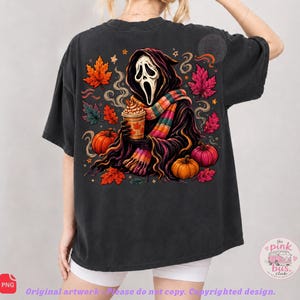 May include: A charcoal black t-shirt featuring a graphic print of a hooded figure with a white mask holding a coffee cup, surrounded by orange and pink autumn leaves, pumpkins, and stars. The design includes a colorful striped scarf and swirls.